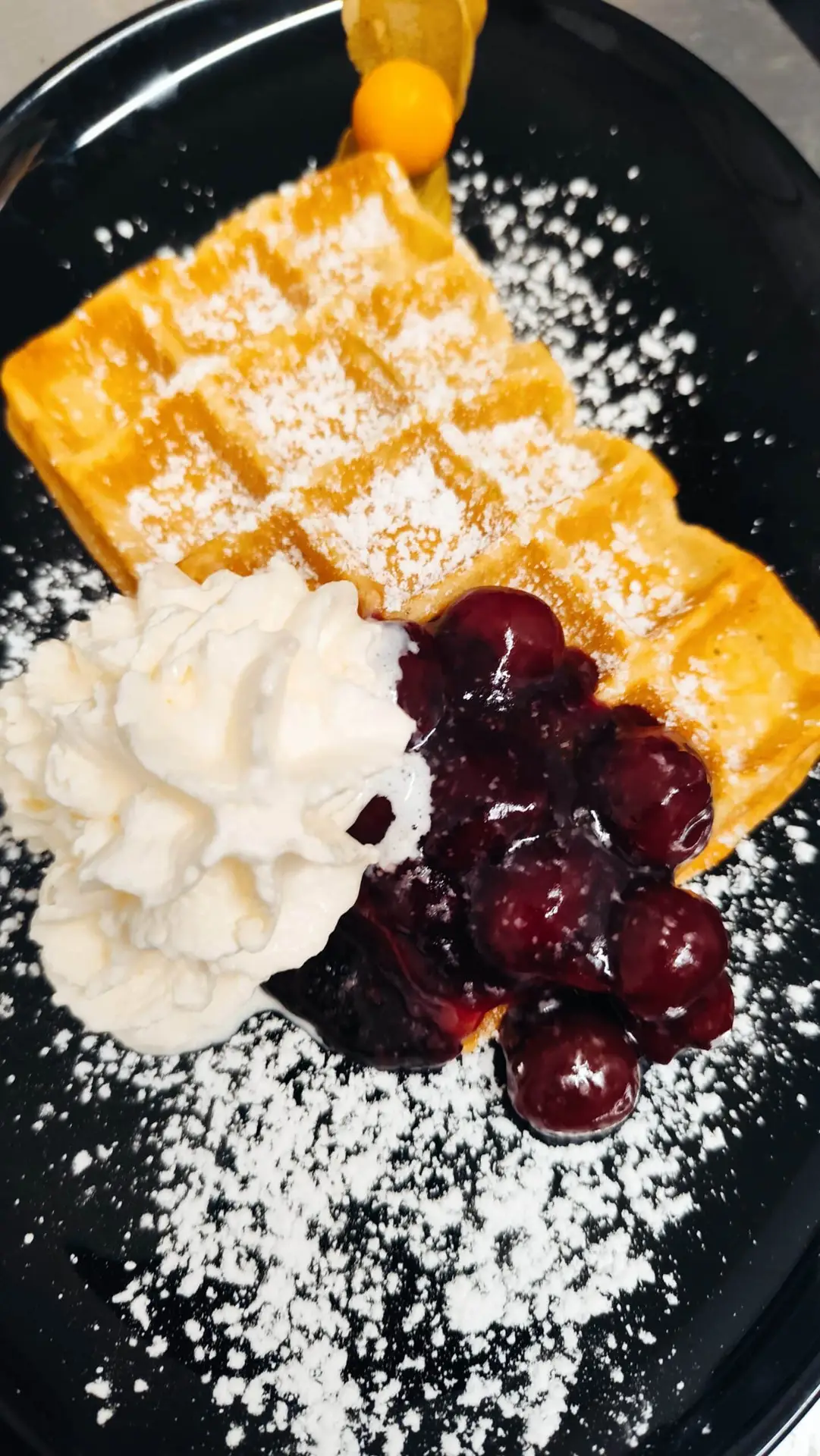 Goldene Waffel mit Sahne Kirschen und Puderzucker auf schwarzem Teller